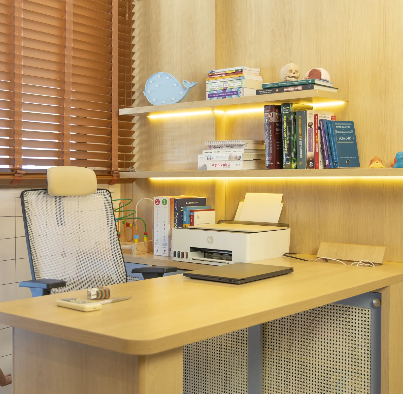Consultório de neuropediatria infantil da Dra. Gabriela Ramos, com mesa, cadeira ergonômica e estante de livros.