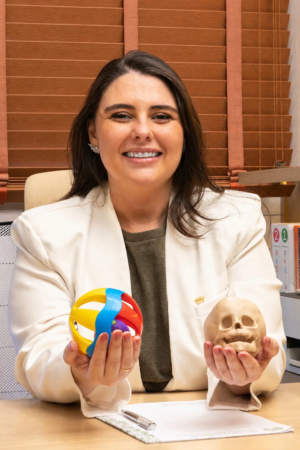 Dra. Gabriela Ramos, neuropediatra, segurando um crânio anatômico e um chocalho com as cores do espectro autista em seu consultório no Rio de Janeiro.